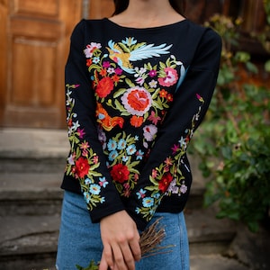 Puede incluir: Una camisa negra de manga larga con bordado floral de colores. El bordado presenta un p&aacute;jaro, flores y hojas. La camisa se combina con una falda vaquera.
