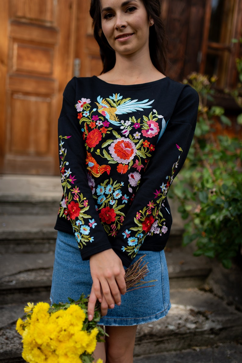 Puede incluir: Una camisa negra de manga larga con bordado floral de colores. El bordado presenta un p&aacute;jaro, flores y hojas. La camisa se combina con una falda vaquera.