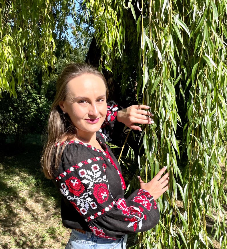Puede incluir: Una mujer con una blusa negra bordada con motivos florales rojos y blancos. Est&aacute; de pie junto a un sauce llor&oacute;n.