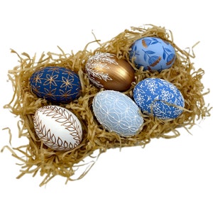 May include: Six decorated Easter eggs in a nest of shredded paper. The eggs are painted in shades of blue and white with gold accents. Some eggs feature floral patterns, while others have geometric designs.