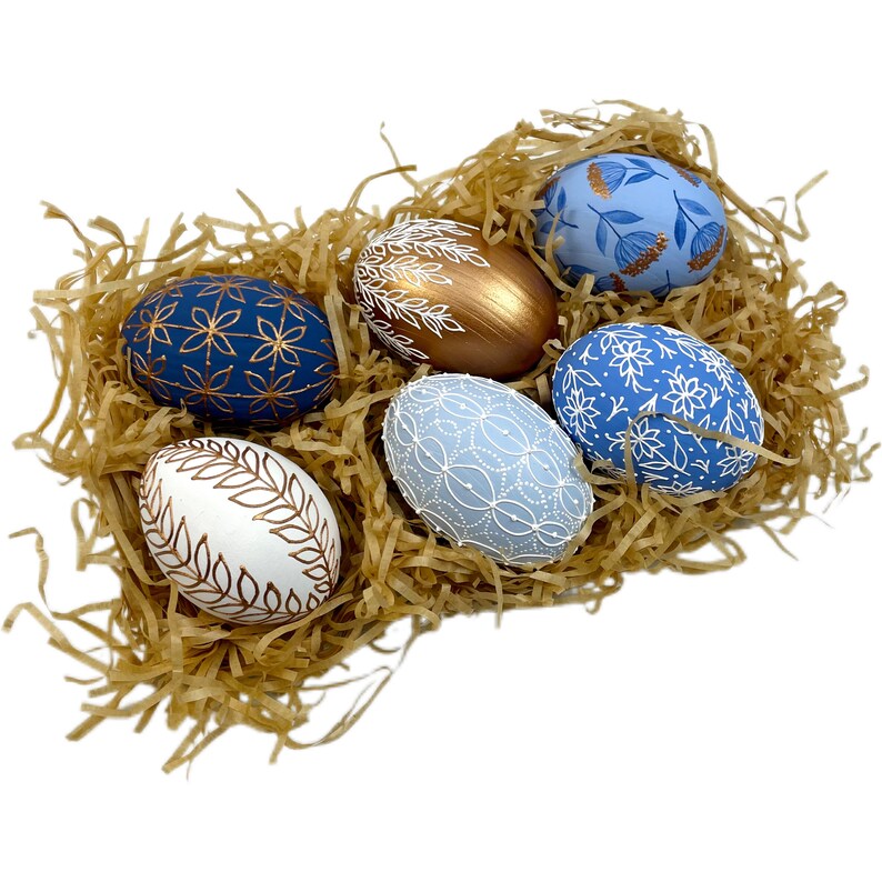 May include: Six decorated Easter eggs in a nest of shredded paper. The eggs are painted in shades of blue and white with gold accents. Some eggs feature floral patterns, while others have geometric designs.