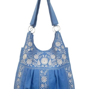Peut inclure: Un sac cabas en toile bleu avec des motifs floraux brod&eacute;s blancs. Le sac a une seule bandouli&egrave;re et un fond pliss&eacute;.