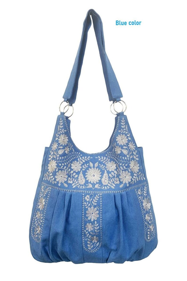 Peut inclure: Un sac cabas en toile bleu avec des motifs floraux brod&eacute;s blancs. Le sac a une seule bandouli&egrave;re et un fond pliss&eacute;.
