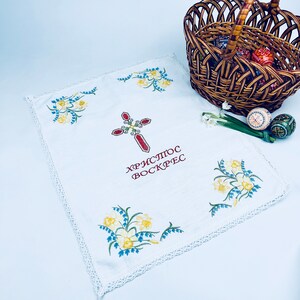 以下が含まれることがあります： 十字架と花の模様が刺繍された白いイースタークロス。布には赤い文字で「キリストは復活した」と書かれています。装飾されたイースターエッグが入った柳のバスケットが背景にあります。