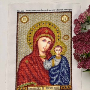 May include: A detailed cross-stitch of an icon featuring a woman and child, with text in Russian. The woman wears a red head covering and holds the child, who is draped in blue. The artwork is framed with gold and brown beads.