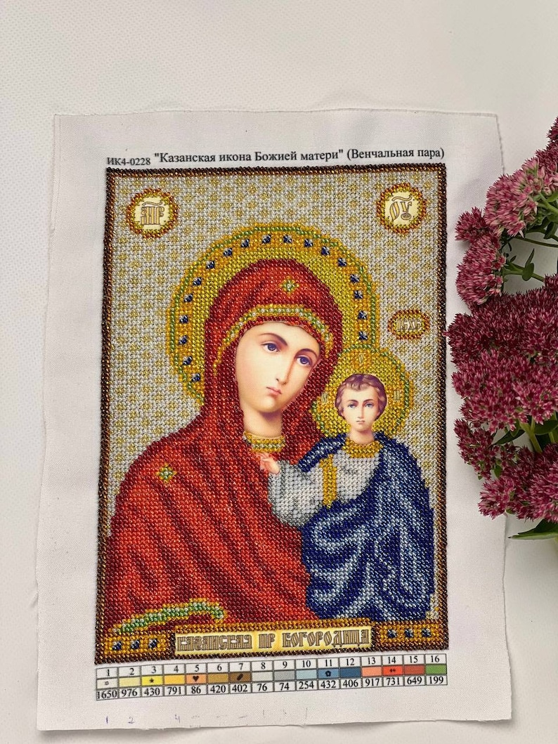 May include: A detailed cross-stitch of an icon featuring a woman and child, with text in Russian. The woman wears a red head covering and holds the child, who is draped in blue. The artwork is framed with gold and brown beads.