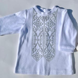 K&ouml;nnte beinhalten: Wei&szlig;es Langarmshirt mit aufwendiger silberner Stickerei auf der Vorderseite. Die Stickerei zeigt ein symmetrisches Blumenmuster. Das Shirt ist aus weichem Stoff und hat einen runden Ausschnitt. Baumwollknospen sind im Hintergrund zu sehen.