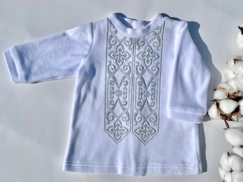 K&ouml;nnte beinhalten: Wei&szlig;es Langarmshirt mit aufwendiger silberner Stickerei auf der Vorderseite. Die Stickerei zeigt ein symmetrisches Blumenmuster. Das Shirt ist aus weichem Stoff und hat einen runden Ausschnitt. Baumwollknospen sind im Hintergrund zu sehen.