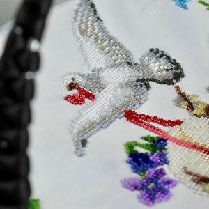May include: A white fabric with a beaded design of a dove in flight, holding a red and white ribbon in its beak. The dove is surrounded by beaded flowers in shades of blue, purple, and green.