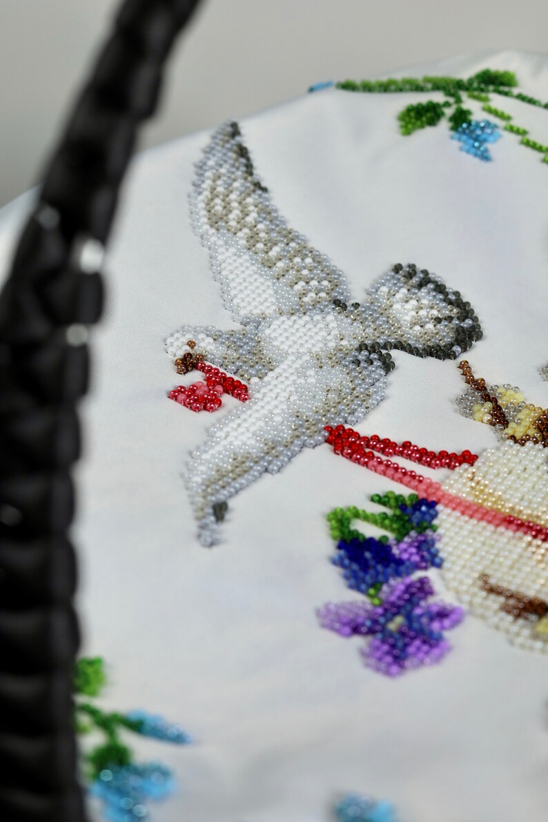 May include: A white fabric with a beaded design of a dove in flight, holding a red and white ribbon in its beak. The dove is surrounded by beaded flowers in shades of blue, purple, and green.