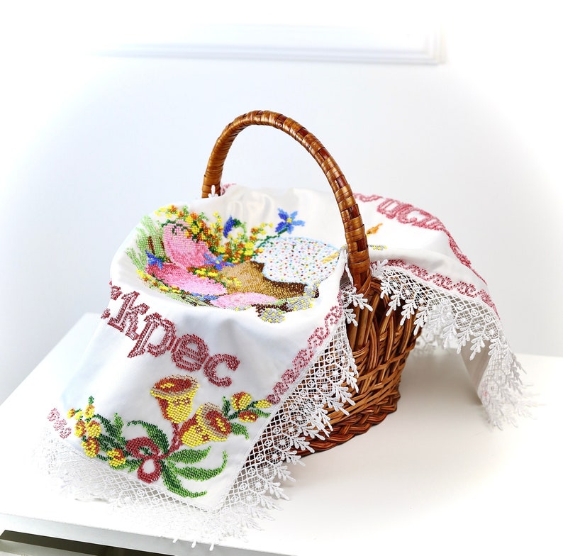 May include: A wicker basket with a handle, containing a white embroidered cloth. The cloth is decorated with colorful floral and religious embroidery, and has a white lace trim. The basket sits on a white surface.