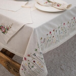 May include: A white linen tablecloth with delicate floral embroidery and a lace trim. The tablecloth is set with a white plate and a folded napkin.