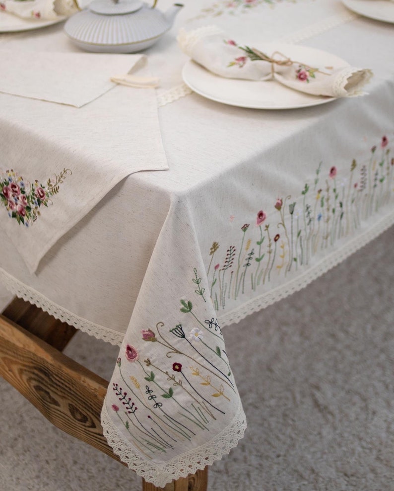 May include: A white linen tablecloth with delicate floral embroidery and a lace trim. The tablecloth is set with a white plate and a folded napkin.
