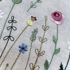 May include: Embroidered floral design on a white fabric background. The design features a variety of flowers and leaves in shades of green, blue, yellow, pink, and red.