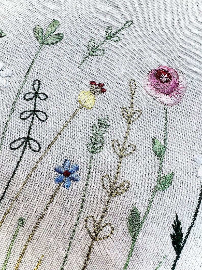 May include: Embroidered floral design on a white fabric background. The design features a variety of flowers and leaves in shades of green, blue, yellow, pink, and red.