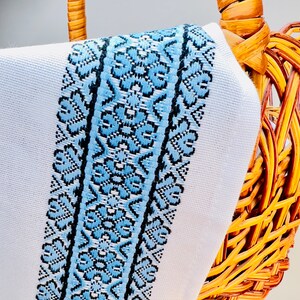 May include: A white fabric with a blue and black embroidered border. The fabric is draped over a wicker basket.
