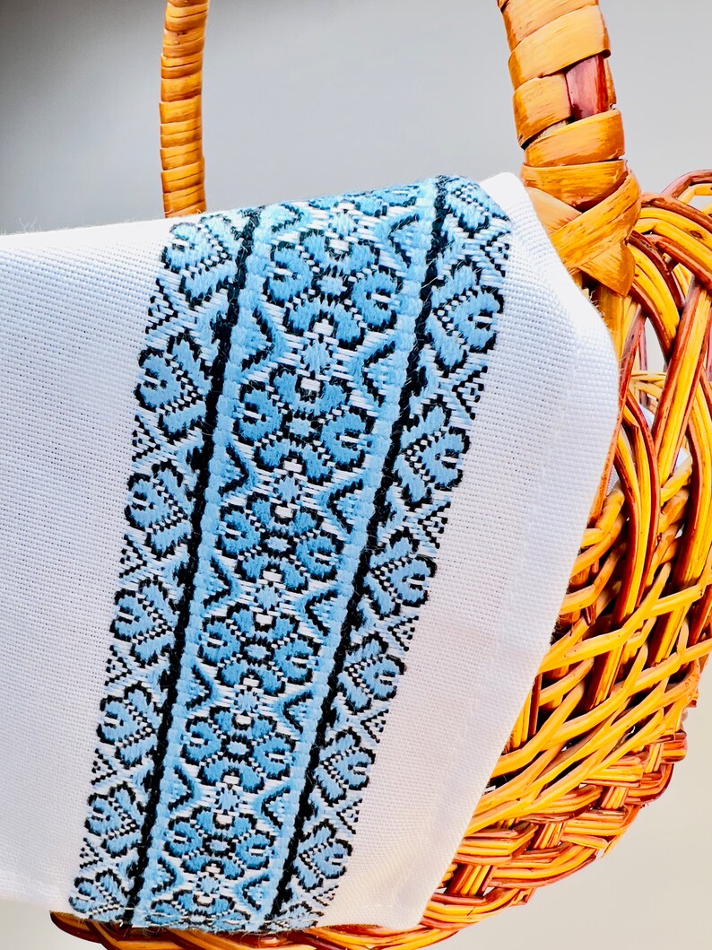 May include: A white fabric with a blue and black embroidered border. The fabric is draped over a wicker basket.