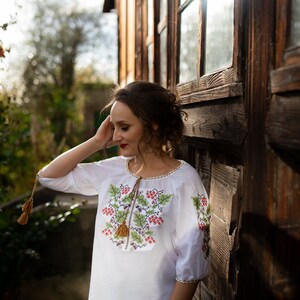 May include: A woman wearing a white embroidered blouse with red and green floral designs and a brown tassel. She is standing next to a wooden building with windows.