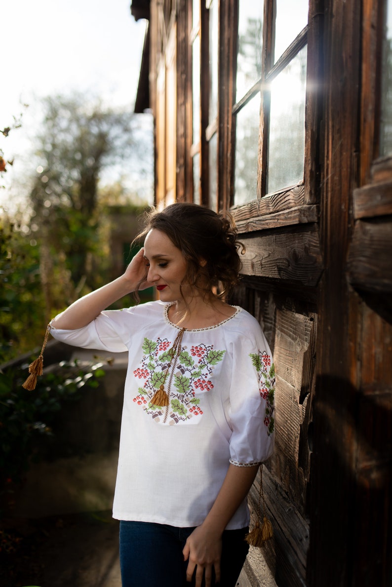 May include: A woman wearing a white embroidered blouse with red and green floral designs and a brown tassel. She is standing next to a wooden building with windows.