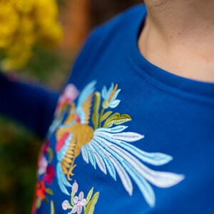 Puede incluir: Una camisa azul con un dise&ntilde;o bordado de colores con un p&aacute;jaro y flores.