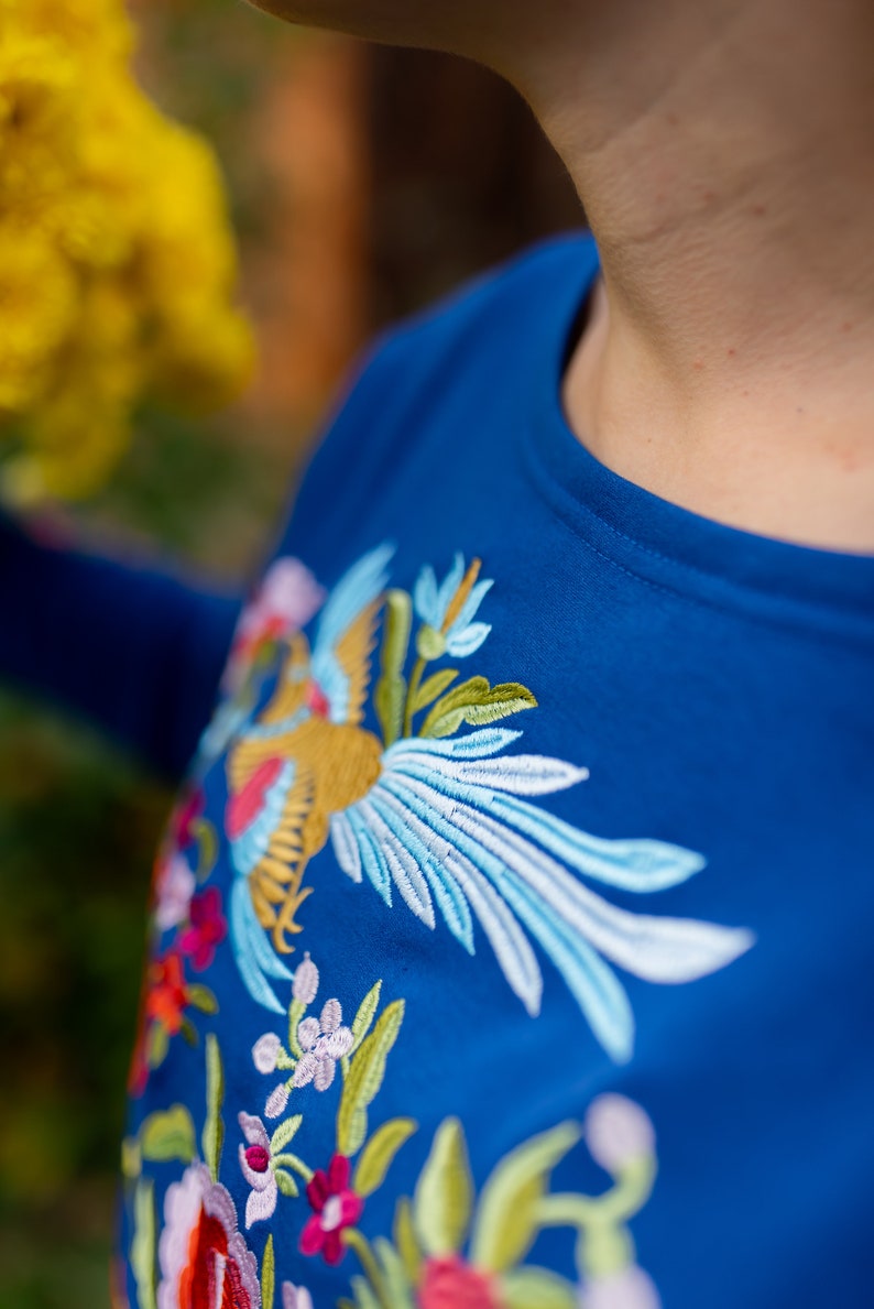 Puede incluir: Una camisa azul con un dise&ntilde;o bordado de colores con un p&aacute;jaro y flores.