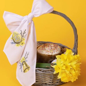K&ouml;nnte beinhalten: Ein Weidenkorb gef&uuml;llt mit Osterleckereien, darunter ein verzierter Kuchen, gelbe Narzissen und ein wei&szlig; besticktes Tuch mit Schleife.