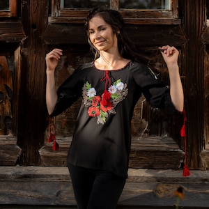 May include: A woman wearing a black embroidered blouse with red and white flowers. The blouse has long sleeves with tassels on the cuffs. She is standing in front of a wooden door.