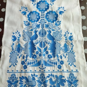 May include: White linen textile with intricate blue embroidery. The design features stylized flowers, birds, and foliage arranged in a symmetrical pattern. The embroidery is detailed and covers a large portion of the fabric, creating a decorative piece.