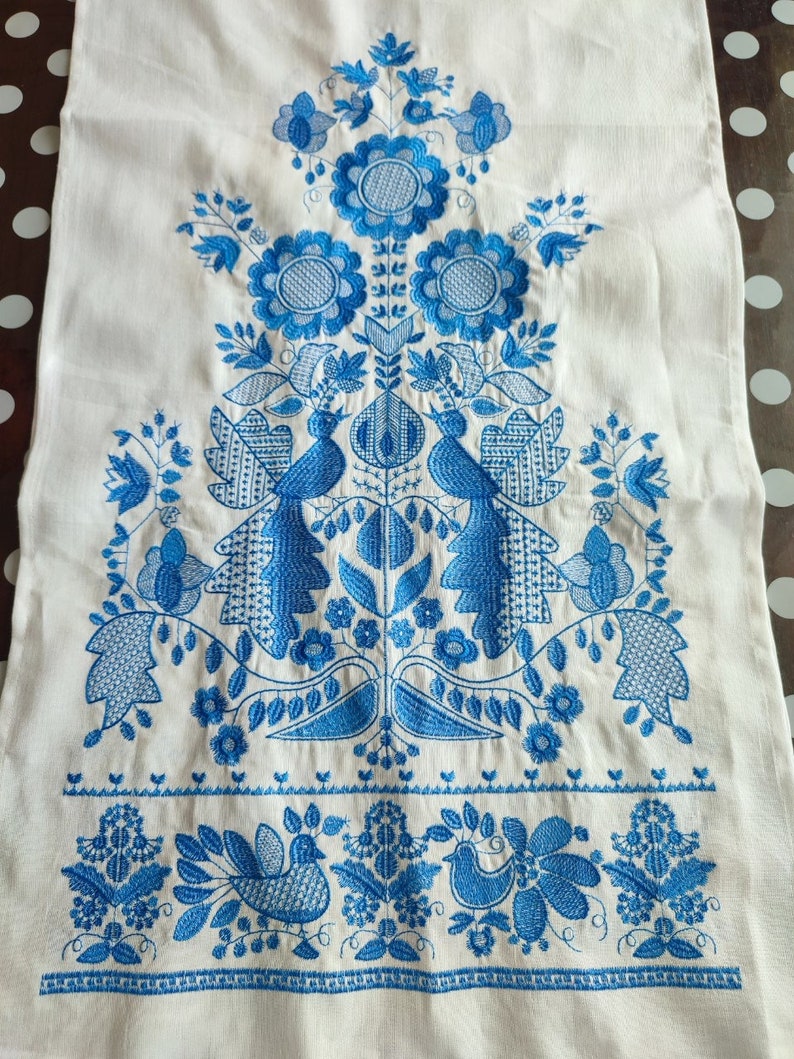 May include: White linen textile with intricate blue embroidery. The design features stylized flowers, birds, and foliage arranged in a symmetrical pattern. The embroidery is detailed and covers a large portion of the fabric, creating a decorative piece.