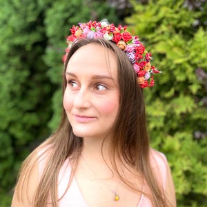 Może przedstawiać: Kobieta w r&oacute;żowej bluzie i koronie kwiatowej wykonanej z czerwonych, r&oacute;żowych i białych kwiat&oacute;w. Nosi srebrny naszyjnik z ż&oacute;łtym kamieniem.