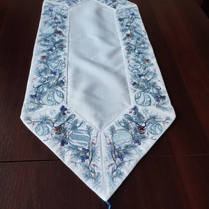 K&ouml;nnte beinhalten: Hellblau-wei&szlig; bestickter Tischl&auml;ufer mit Weihnachtsmotiv. Der L&auml;ufer zeigt blaue Ornamente, Tannennadeln und Beeren. Unten h&auml;ngt eine blaue Quaste. Der L&auml;ufer ist etwa 100 cm lang.