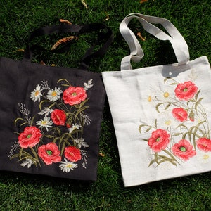Op de afbeelding: Twee tote bags met geborduurde klaprozen. E&eacute;n tas is zwart met witte bloemen en de andere is wit met groene bloemen.