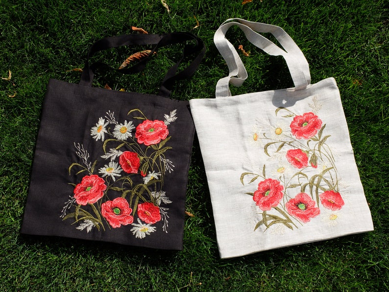 Op de afbeelding: Twee tote bags met geborduurde klaprozen. E&eacute;n tas is zwart met witte bloemen en de andere is wit met groene bloemen.