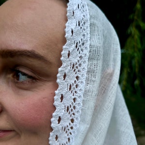 May include: A white head covering with a delicate, openwork lace trim. The fabric appears to be a lightweight linen or cotton, draped over the head. The lace trim features a repeating floral pattern, adding a touch of elegance.