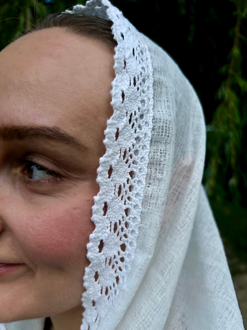 May include: A white head covering with a delicate, openwork lace trim. The fabric appears to be a lightweight linen or cotton, draped over the head. The lace trim features a repeating floral pattern, adding a touch of elegance.