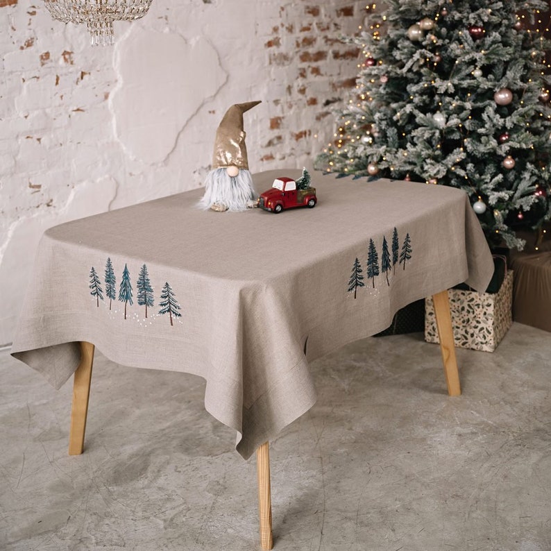 May include: A beige linen tablecloth with embroidered Christmas trees. A small red truck carrying a Christmas tree and a gnome figurine are on the table. The table has wooden legs and is set in front of a Christmas tree.
