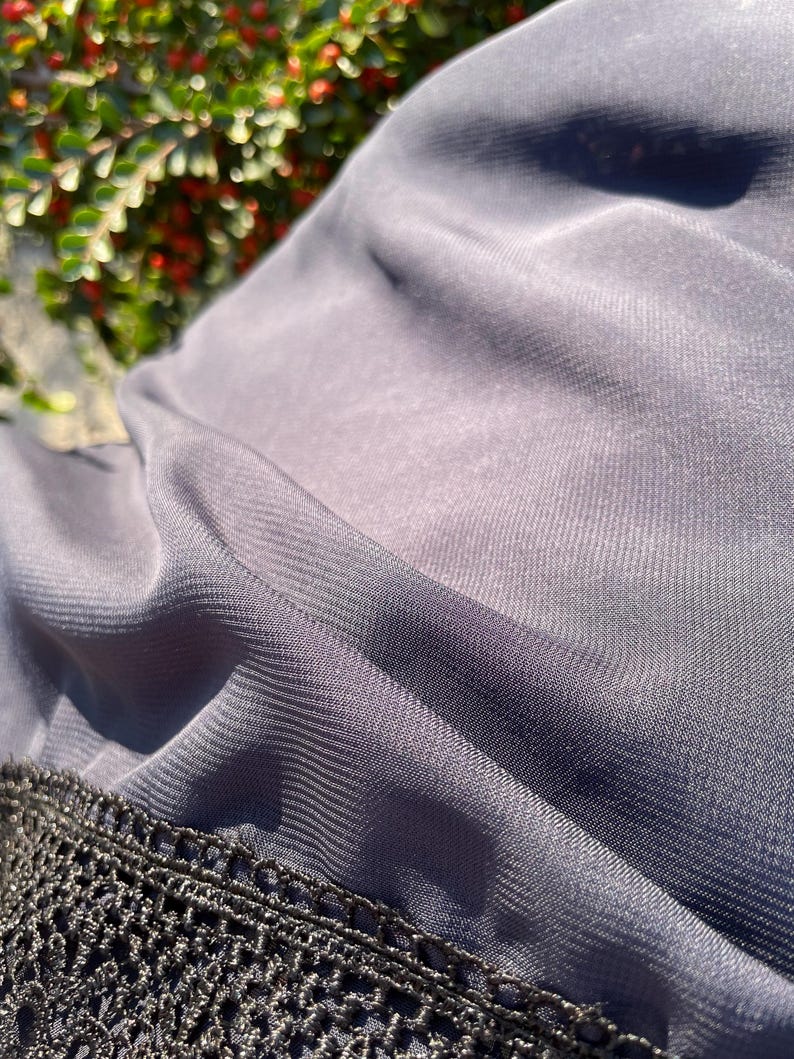 May include: A close-up of a dark gray sheer fabric with a black crocheted trim. The fabric is soft and flowing, with a subtle sheen.