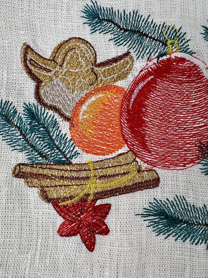 May include: Embroidered Christmas design featuring a red ornament, orange sphere, and gold angel. Green pine needles and cinnamon sticks with a red star are also included. The design is on a white fabric background.