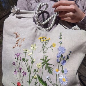 Op de afbeelding: Een lichtgrijze canvas tote bag met een wit touw handvat. De tas is geborduurd met verschillende kleurrijke wilde bloemen, waaronder blauwe, paarse, gele en witte.