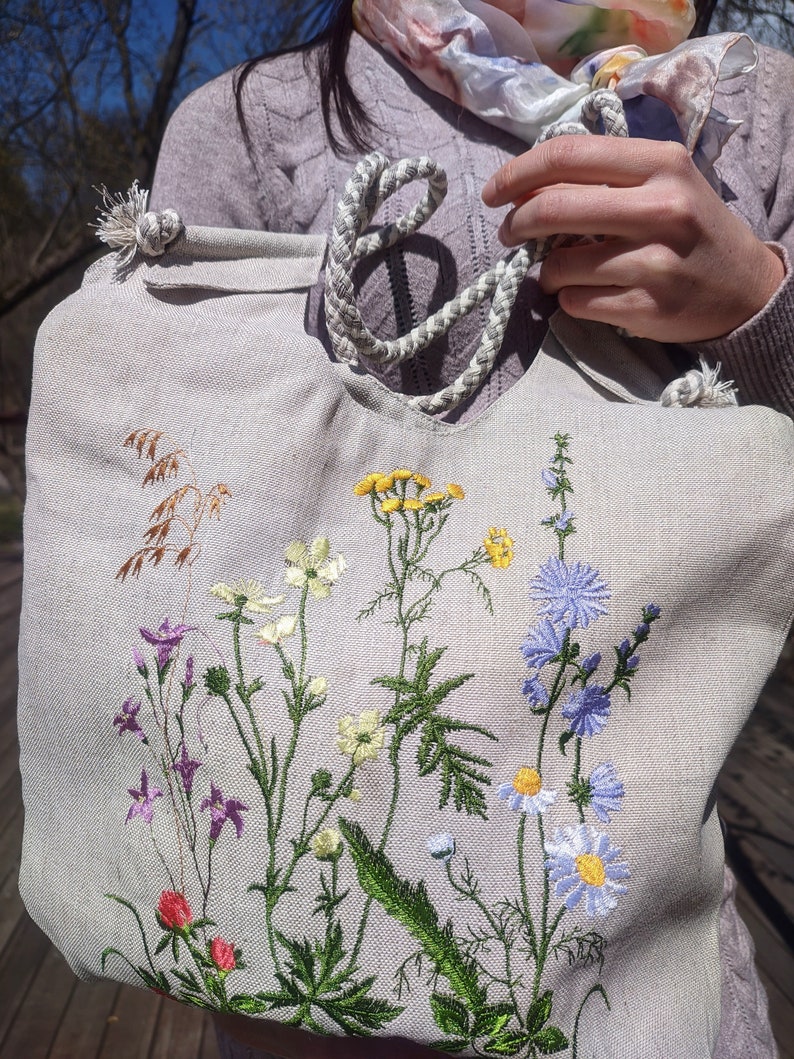 Op de afbeelding: Een lichtgrijze canvas tote bag met een wit touw handvat. De tas is geborduurd met verschillende kleurrijke wilde bloemen, waaronder blauwe, paarse, gele en witte.