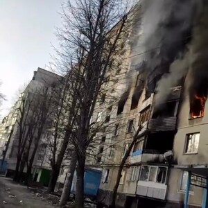 K&ouml;nnte beinhalten: Ein mehrst&ouml;ckiges Wohnhaus mit Feuer und Rauch, der aus mehreren Fenstern quillt. Die Fassade des Geb&auml;udes ist grau, und kahle B&auml;ume s&auml;umen die Stra&szlig;e. Der Himmel ist bedeckt.
