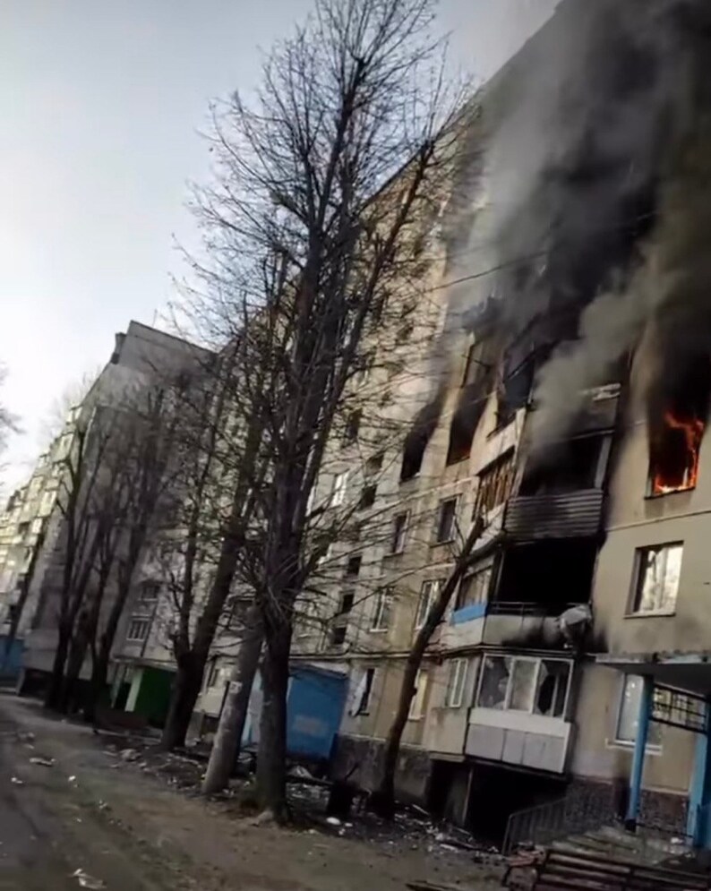 K&ouml;nnte beinhalten: Ein mehrst&ouml;ckiges Wohnhaus mit Feuer und Rauch, der aus mehreren Fenstern quillt. Die Fassade des Geb&auml;udes ist grau, und kahle B&auml;ume s&auml;umen die Stra&szlig;e. Der Himmel ist bedeckt.