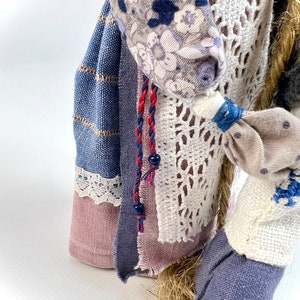 May include: A handmade rag doll with a blue and white striped skirt, a pink and white floral print top, and a white crocheted shawl. The doll has a long, braided hair and is wearing a red and blue braided cord with black beads.