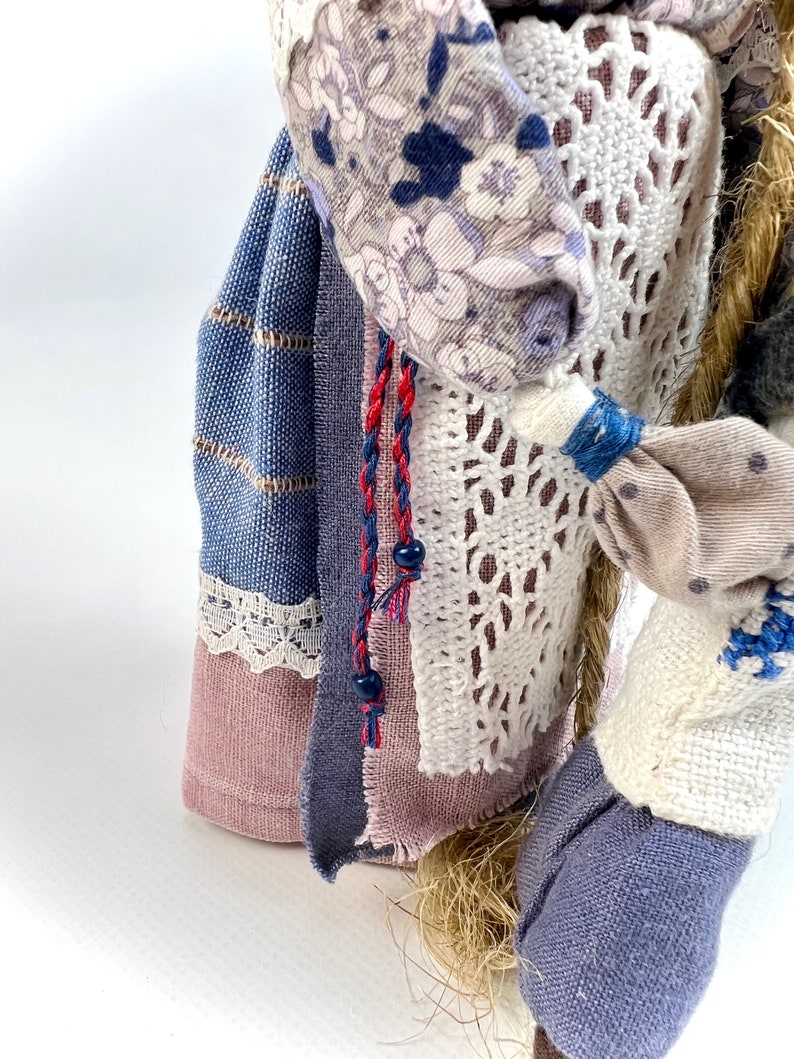 May include: A handmade rag doll with a blue and white striped skirt, a pink and white floral print top, and a white crocheted shawl. The doll has a long, braided hair and is wearing a red and blue braided cord with black beads.