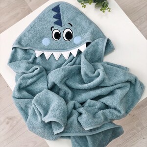 May include: A blue hooded towel with a shark face embroidered on the hood. The towel is made of soft, absorbent material and is perfect for drying a child after bath time.