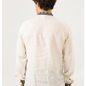 May include: Cream-colored long-sleeve shirt with black and white embroidered details on the collar and cuffs. The shirt is made of a lightweight fabric, likely linen or a similar material. The back view shows the shirt's design and texture.