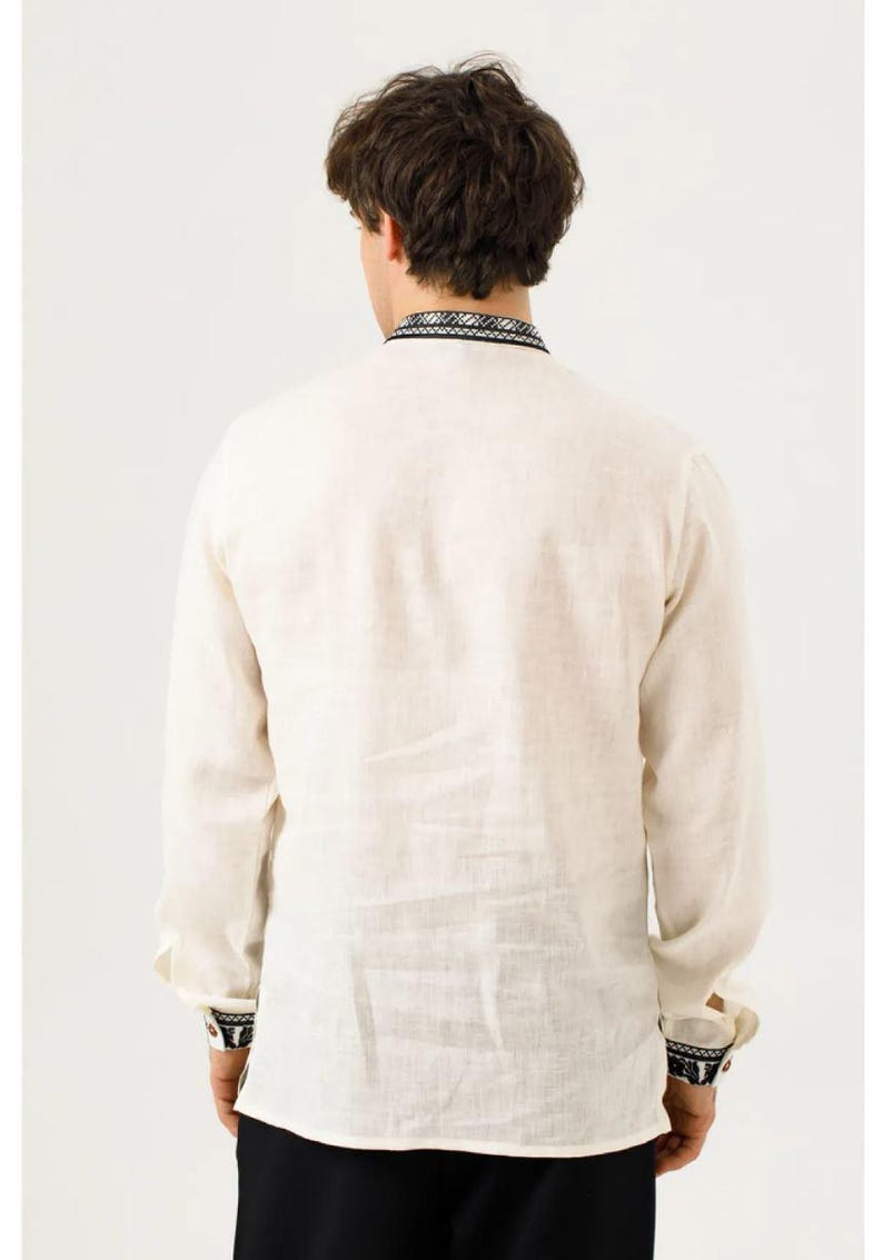 May include: Cream-colored long-sleeve shirt with black and white embroidered details on the collar and cuffs. The shirt is made of a lightweight fabric, likely linen or a similar material. The back view shows the shirt's design and texture.