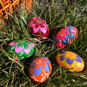 May include: A collection of hand-painted Easter eggs with floral designs in various colors, including pink, green, orange, and yellow. The eggs are nestled in green grass, with a wicker basket partially visible in the background.