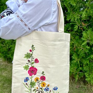 以下が含まれることがあります： 白いキャンバス地のトートバッグに、花柄の刺繍が施されています。刺繍には、ピンク、赤、黄色、青の花がデザインされています。