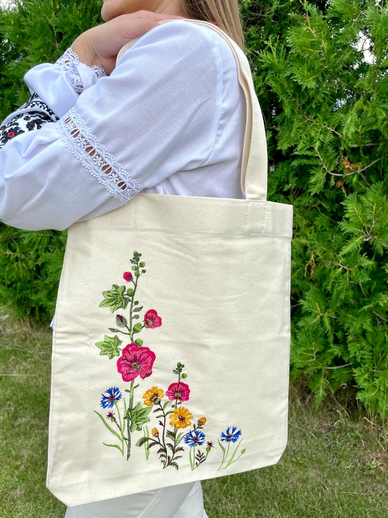 以下が含まれることがあります： 白いキャンバス地のトートバッグに、花柄の刺繍が施されています。刺繍には、ピンク、赤、黄色、青の花がデザインされています。